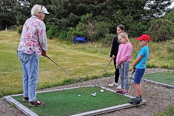 Children standing watching as a golf lesson is provided by an older woman